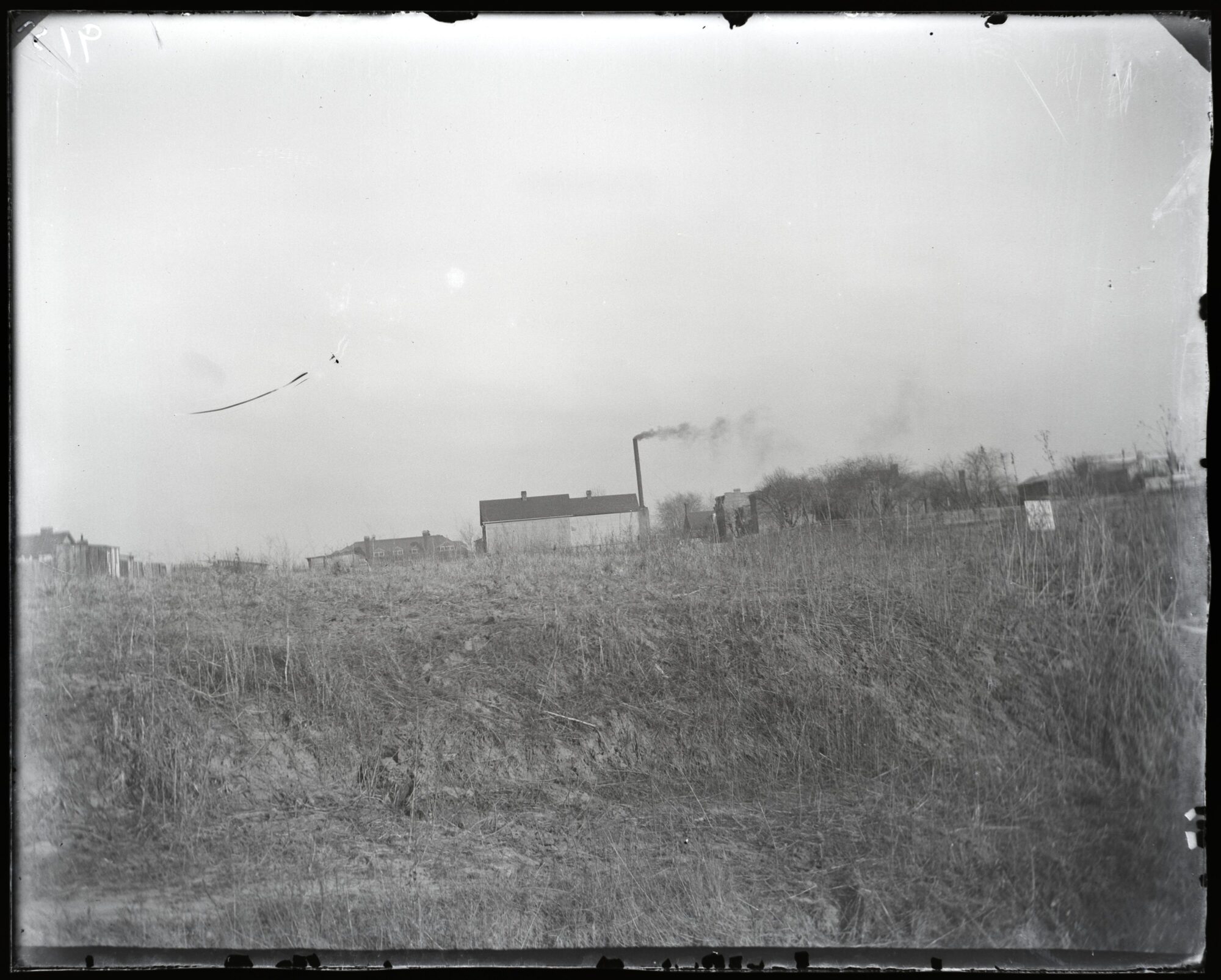 Black and white photograph of the House of Refuge in St. Louis. At center is an unadorned building with a smokestack. Additional buildings are blurry but visible in the background. The foreground is an overgrown grassy expanse.